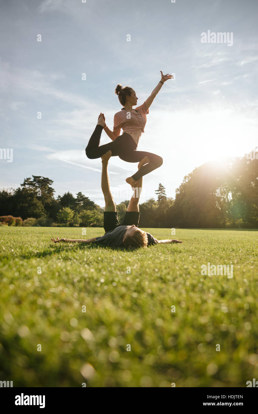 Vertical shot of healthy young couple doing acrobatic yoga on grass. Man and woman exercising in pair at the park. Stock Photo