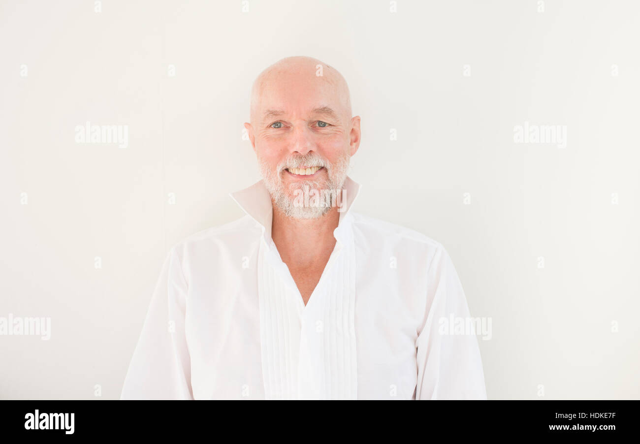 Smiling old man in casual dress looking at camera. Standing in front of white wall in his home. Showing cheerful and positive facial expression. Stock Photo