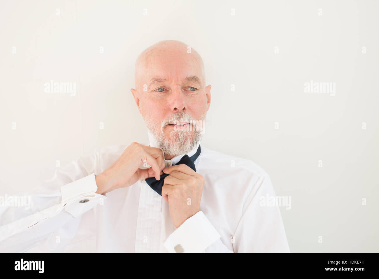 Old man putting on black bowtie and white shirt at home. Lifestyle moment of active retirement and preparing for wedding party or elegant occasion. Stock Photo