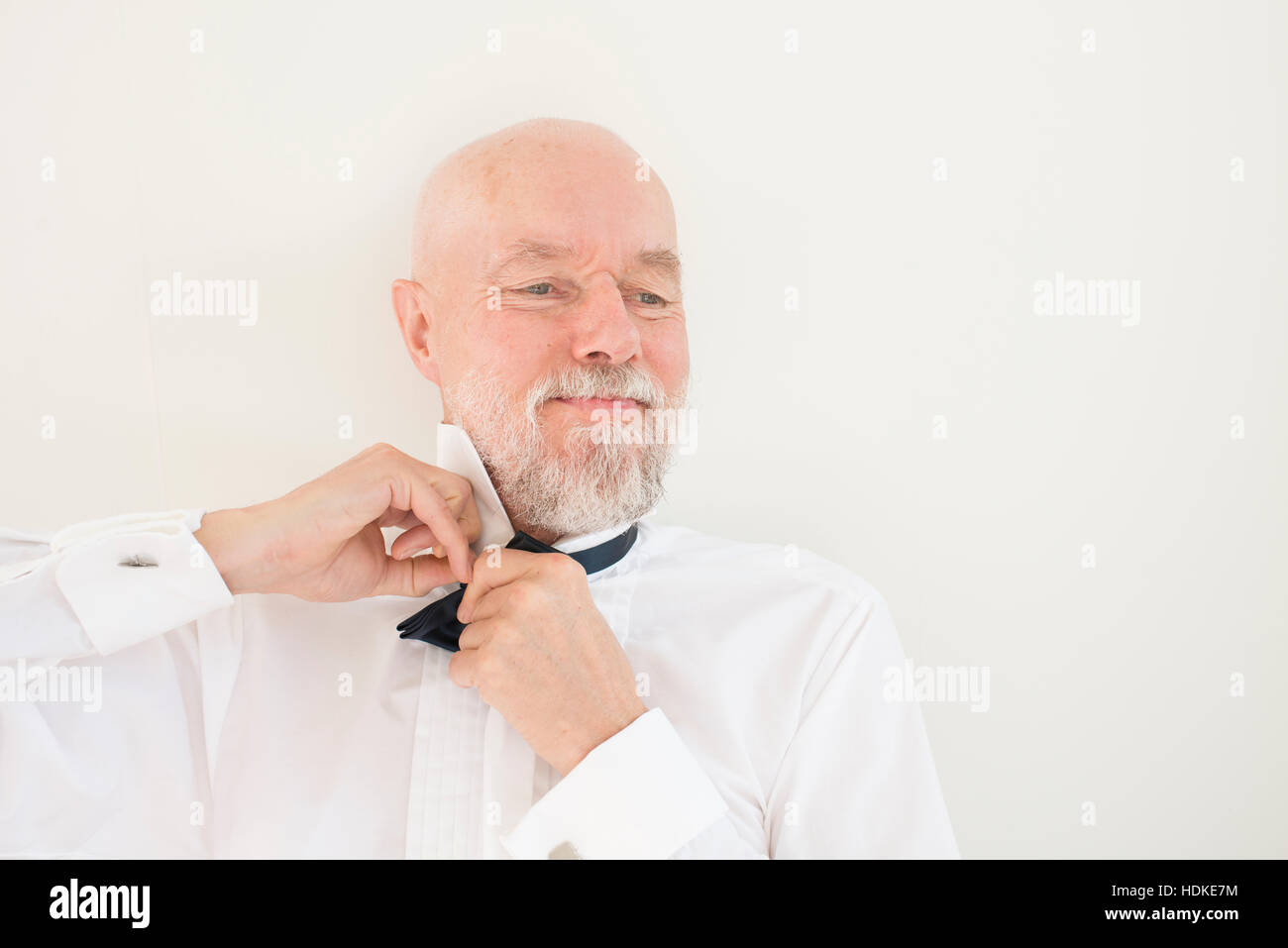 Old man putting on black bowtie and white shirt at home. Lifestyle moment of active retirement and preparing for wedding party or elegant occasion. Stock Photo