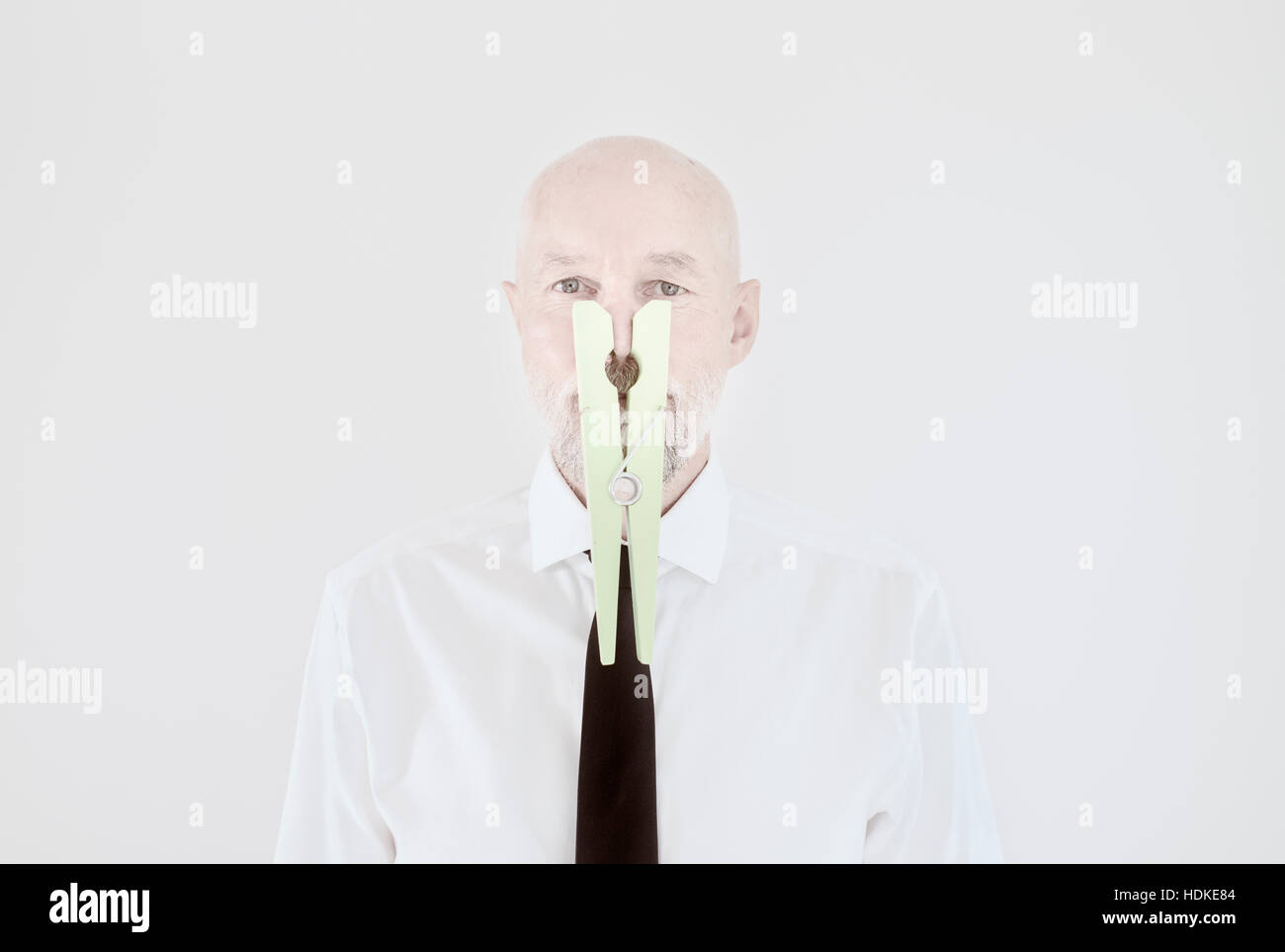 Old man with big clothespin covering his nose. Humorous portrait of elderly male. Concept of bad smell, absurd behaviour and contrast between serious Stock Photo