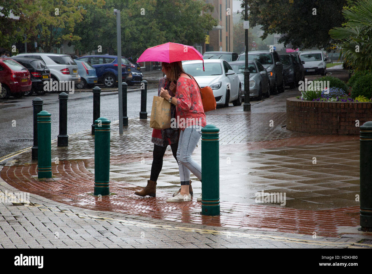 Two women share a pink umbrella while crossing a wet pavement in Crawley,Sussex, UK. Stock Photo