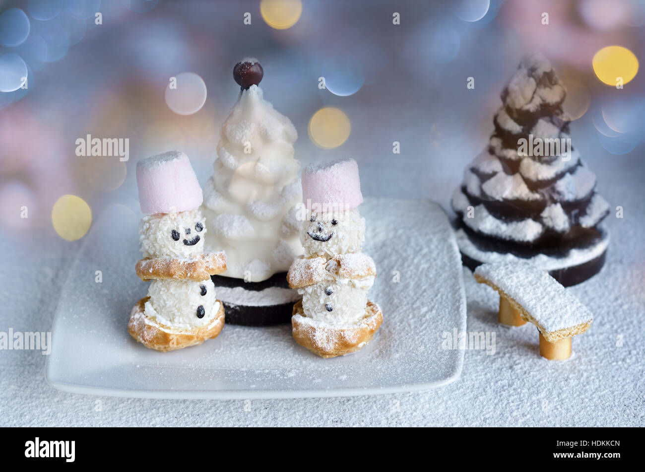 Little cakes snowmen and chocolate trees in powdered sugar. On the background of colorful bokeh. Stock Photo