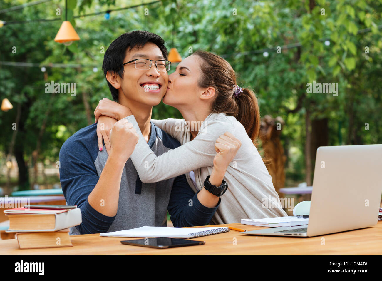 Cute young woman sitting and kissing cheerful asian young man in glasses outdoors Stock Photo