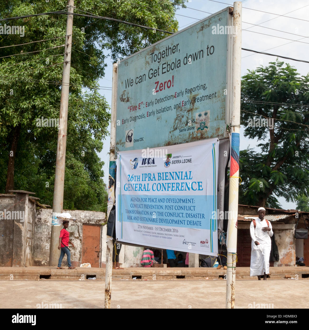 A weathered billboard in Freetown promotes the "five Es" against Ebola – early detection, reporting, diagnosis, isolation and treatment. Below it hangs a banner for the International Peace Research Association's (IPRA) 26th general conference. Western Area, Sierra Leone Stock Photo