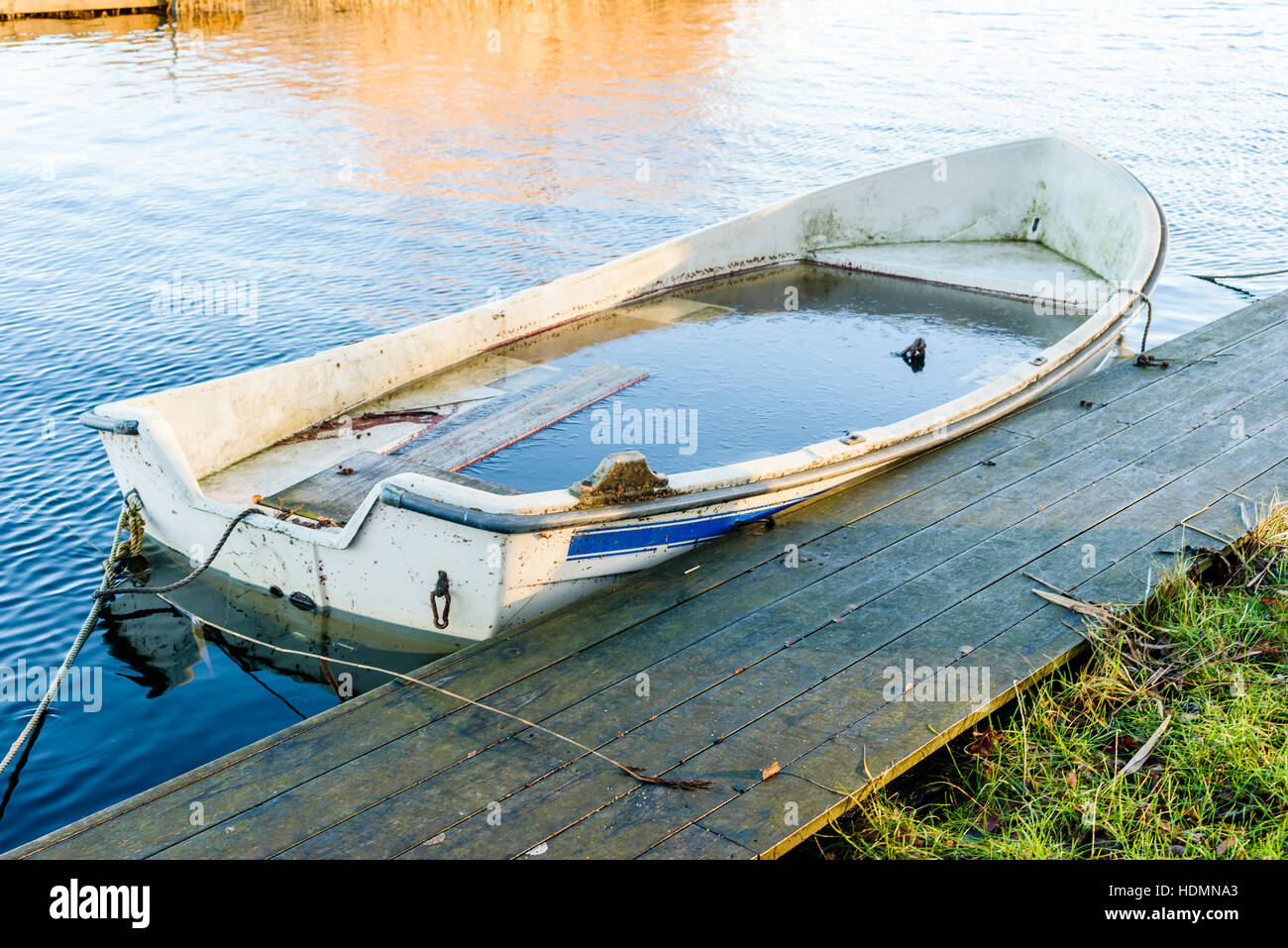 Small water filled rowboat moored beside a narrow wooden pier on a cold winter morning. Ice inside the boat. Stock Photo