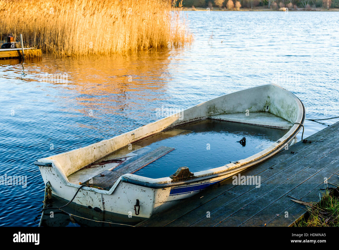 Small water filled rowboat moored beside a narrow wooden pier on a cold winter morning. Ice inside the boat. Stock Photo
