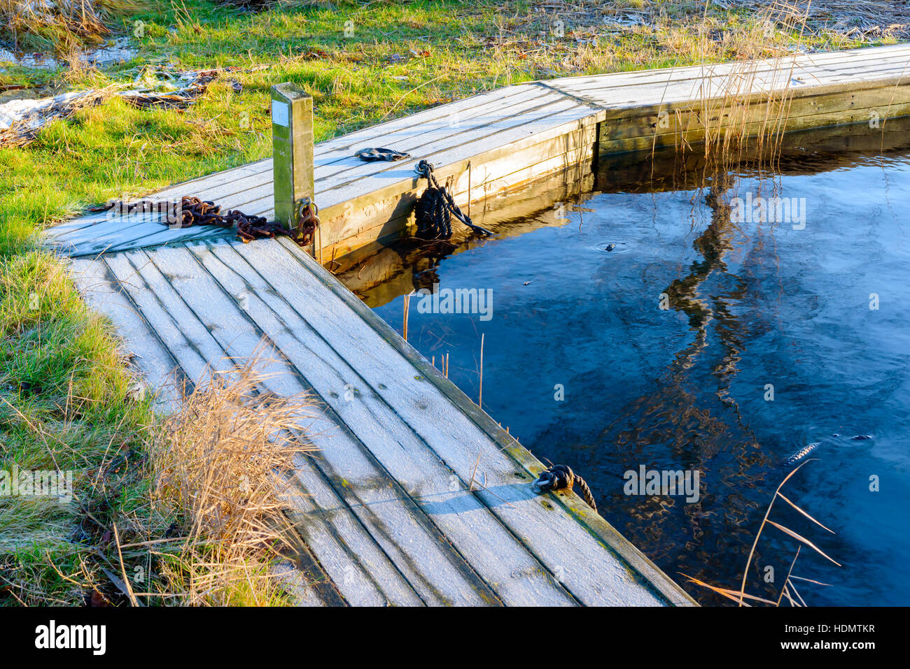 Narrow and small wooden pier covered in frost. Chain and rope at the pier but no boat. Chilly winter morning with calm water. Stock Photo
