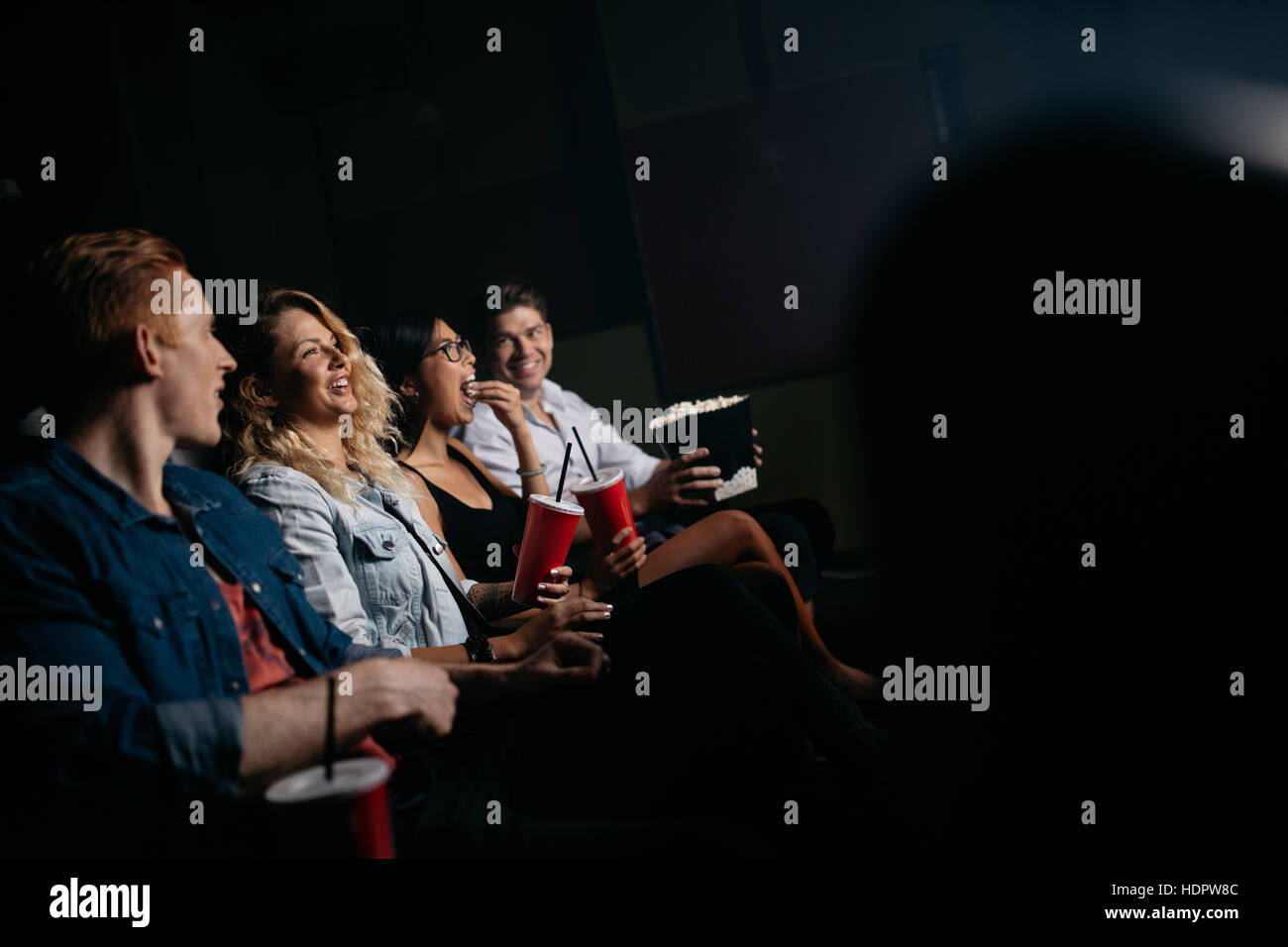 Group of friends sitting in multiplex movie theater with popcorn and drinks. Young people watching movie in cinema. Stock Photo