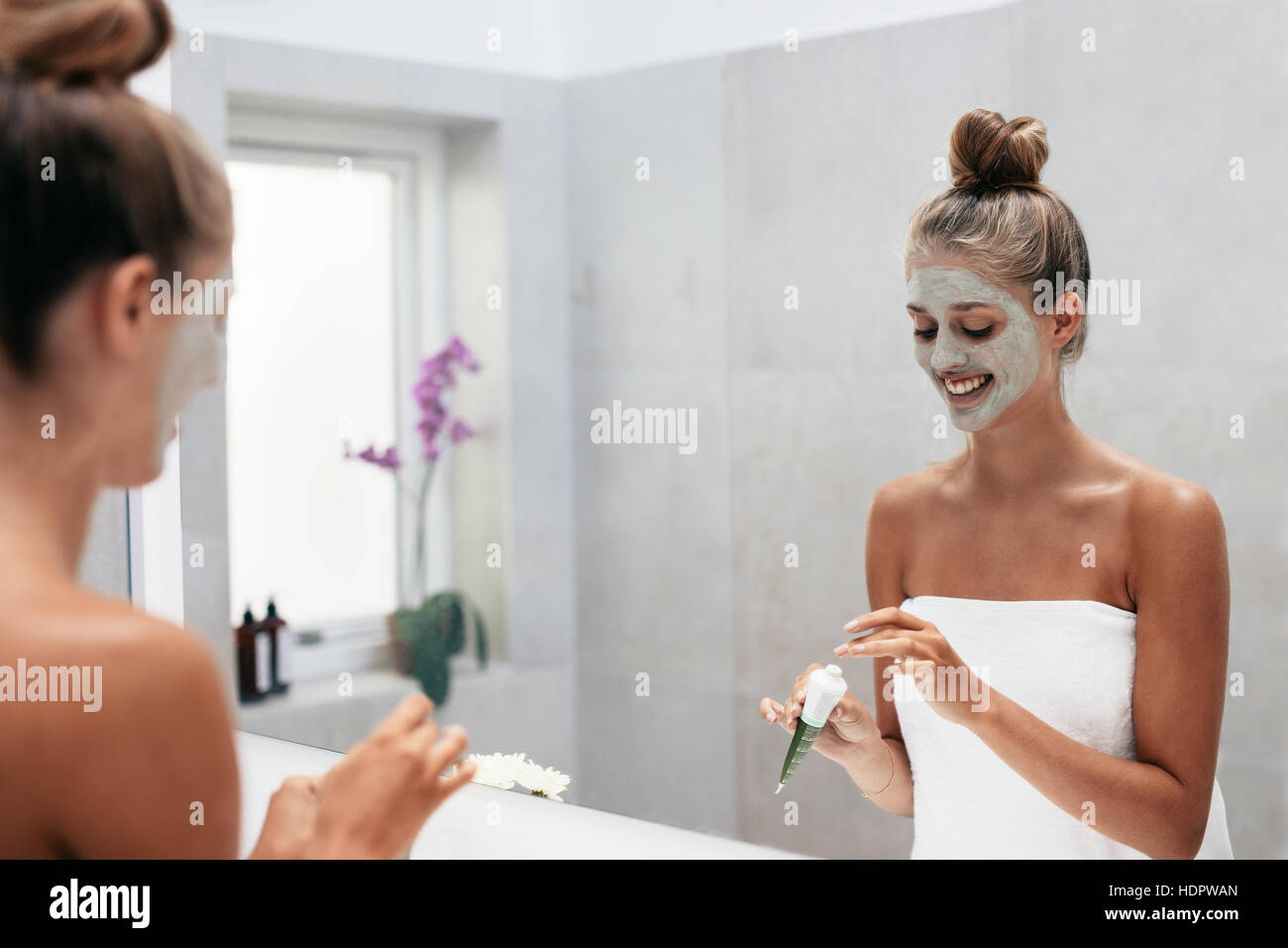 Happy young woman applying face mask in bathroom and smiling. Beautiful female in front of mirror doing beauty treatment. Stock Photo