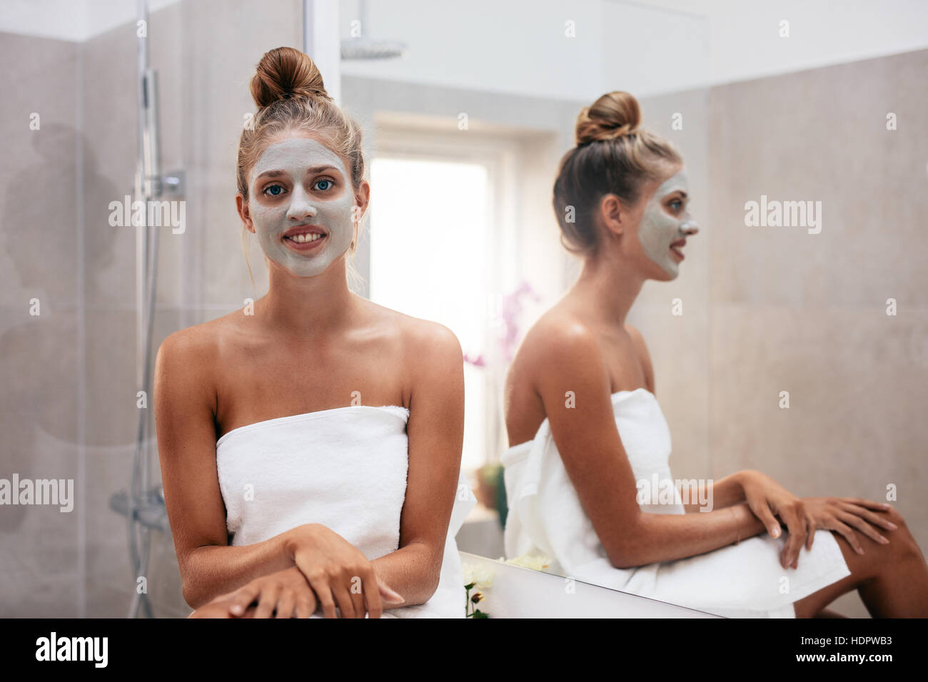 Beautiful young woman with facial mask sitting in bathroom and smiling. Stock Photo