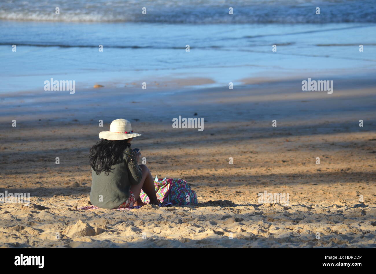 Young woman in hat sitting on the beach and watching her mobile phone ...