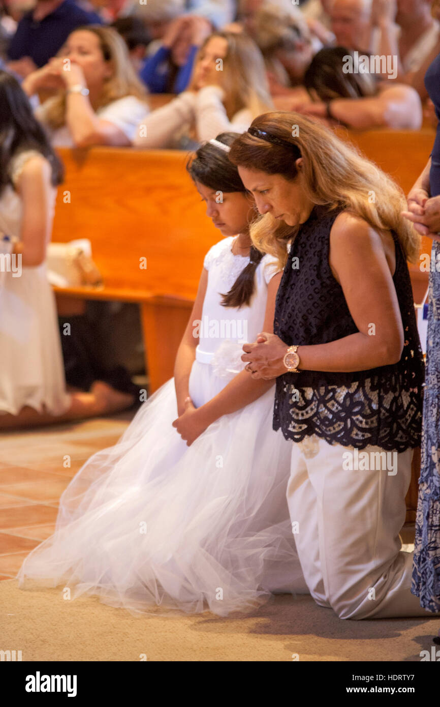 A Hispanic mother and daughter pray during First Communion mass at a Laguna Niguel, CA, Catholic church. Note formal dress on girl and parishioners praying in background. Stock Photo