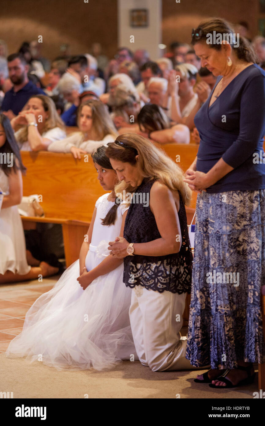 A Hispanic mother and daughter pray during First Communion mass at a Laguna Niguel, CA, Catholic church. Note formal dress on girl and parishioners praying in background. Stock Photo