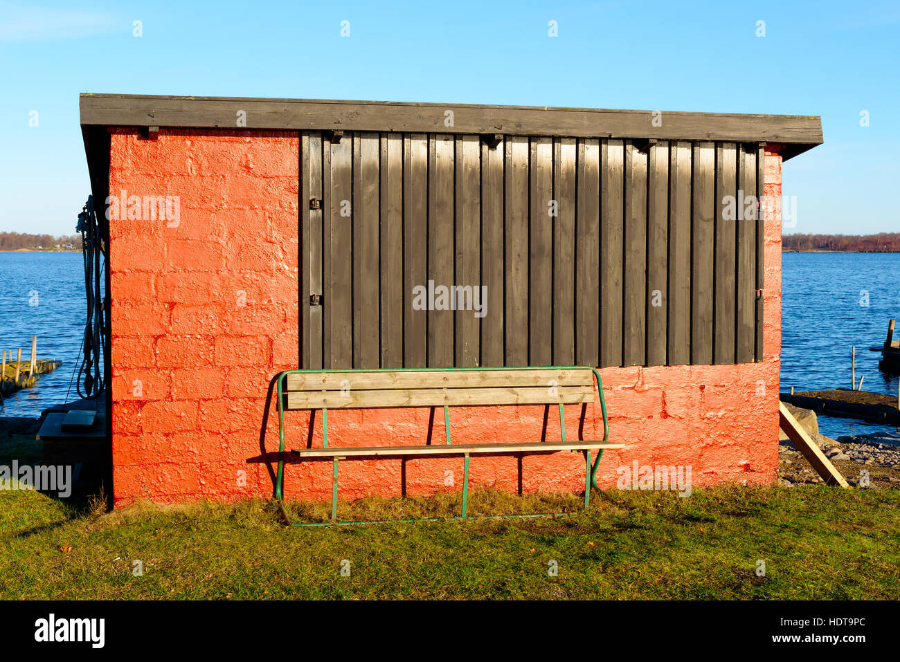 Empty green bench outside a small red storage shed with large wooden hatch. Surrounding coastline visible at sides. Copy space above bench. Stock Photo