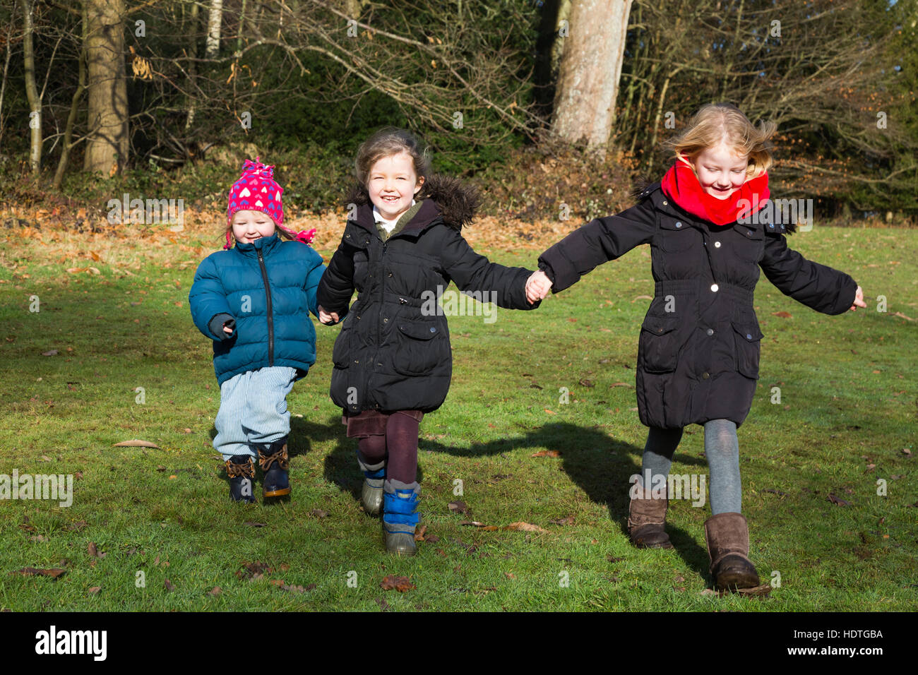 Three sisters: girls age 4, 2 & 6 years, four, two, and six year old, running run play in grass park / parkland / UK autumn. Stock Photo