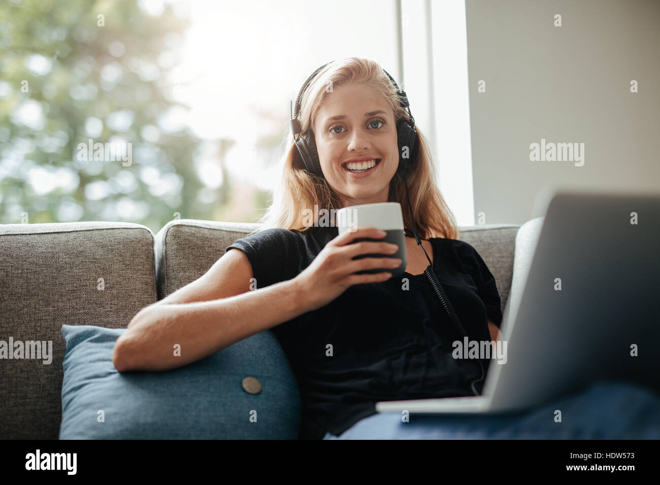 Smiling young woman with a cup of coffee relaxing on sofa with laptop ...