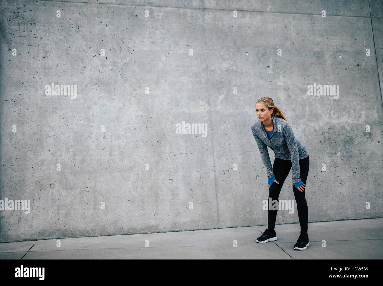 Female runner standing bent over and catching her breath after a running session in city. Young sports woman taking break after a run. Stock Photo