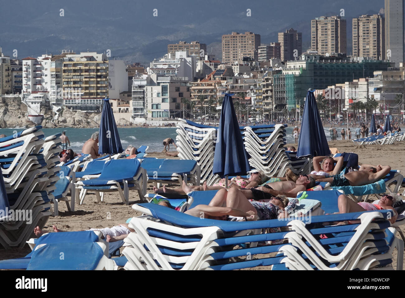 Crowded beach scene with sunbeds and people relaxing or exercising on the shoreline with skyscrapers in back Stock Photo