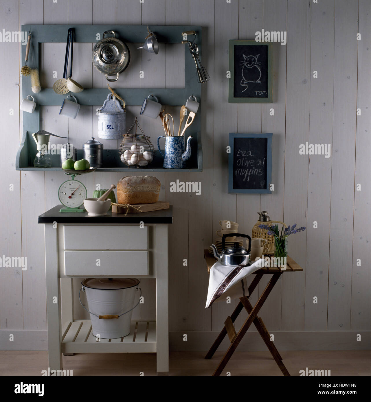 Kitchen utensils on gray painted shelving above a painted butcher's block and a wooden stool with a kettle Stock Photo