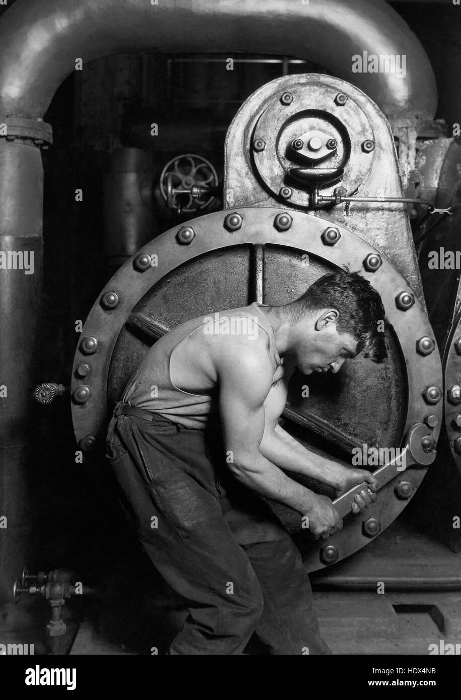 Lewis Hine's 1920 Power house mechanic working on steam pump, one of his 'work portraits', shows a working class American in an industrial setting. The carefully posed subject, a young man with wrench in hand, is hunched over, surrounded by the machinery that defines his job. But while constrained by the machinery (almost a metal womb), the man is straining against it--muscles taut, with a determined look--in an iconic representation of masculinity. Photograph By Lewis W. Hine. Stock Photo