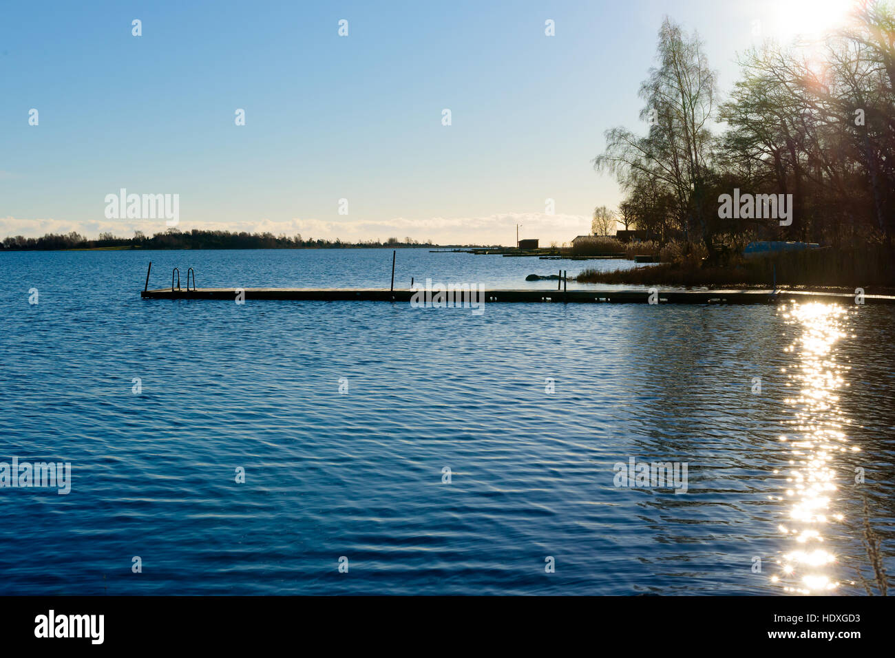 Backlit swimming pier in calm water. Sun and reflections visible over some trees. Stock Photo