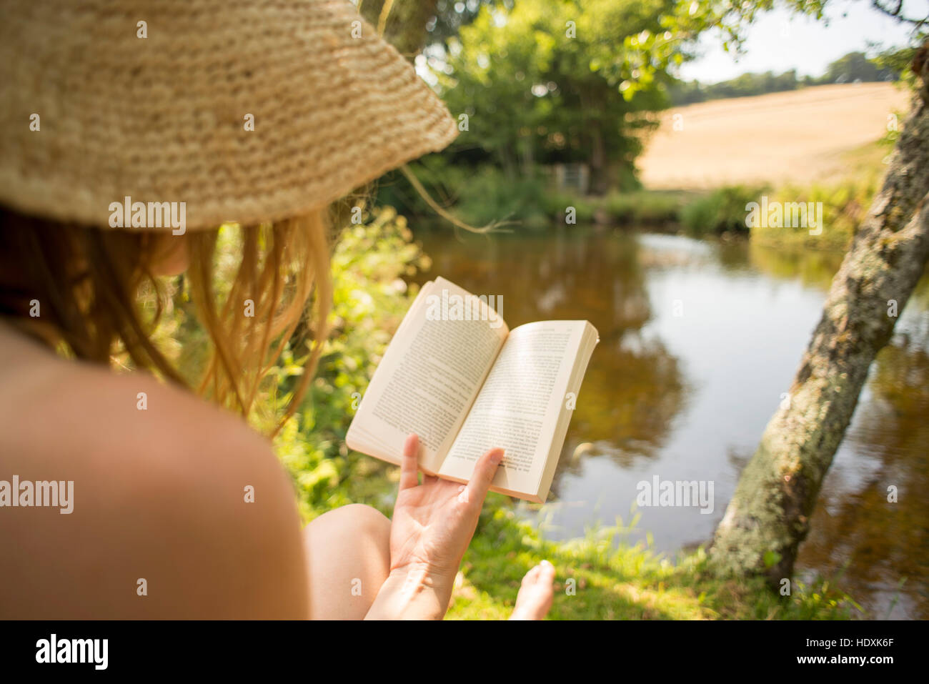 Woman wearing a hat reading a book on holiday in English countryside by a river Devon England Stock Photo