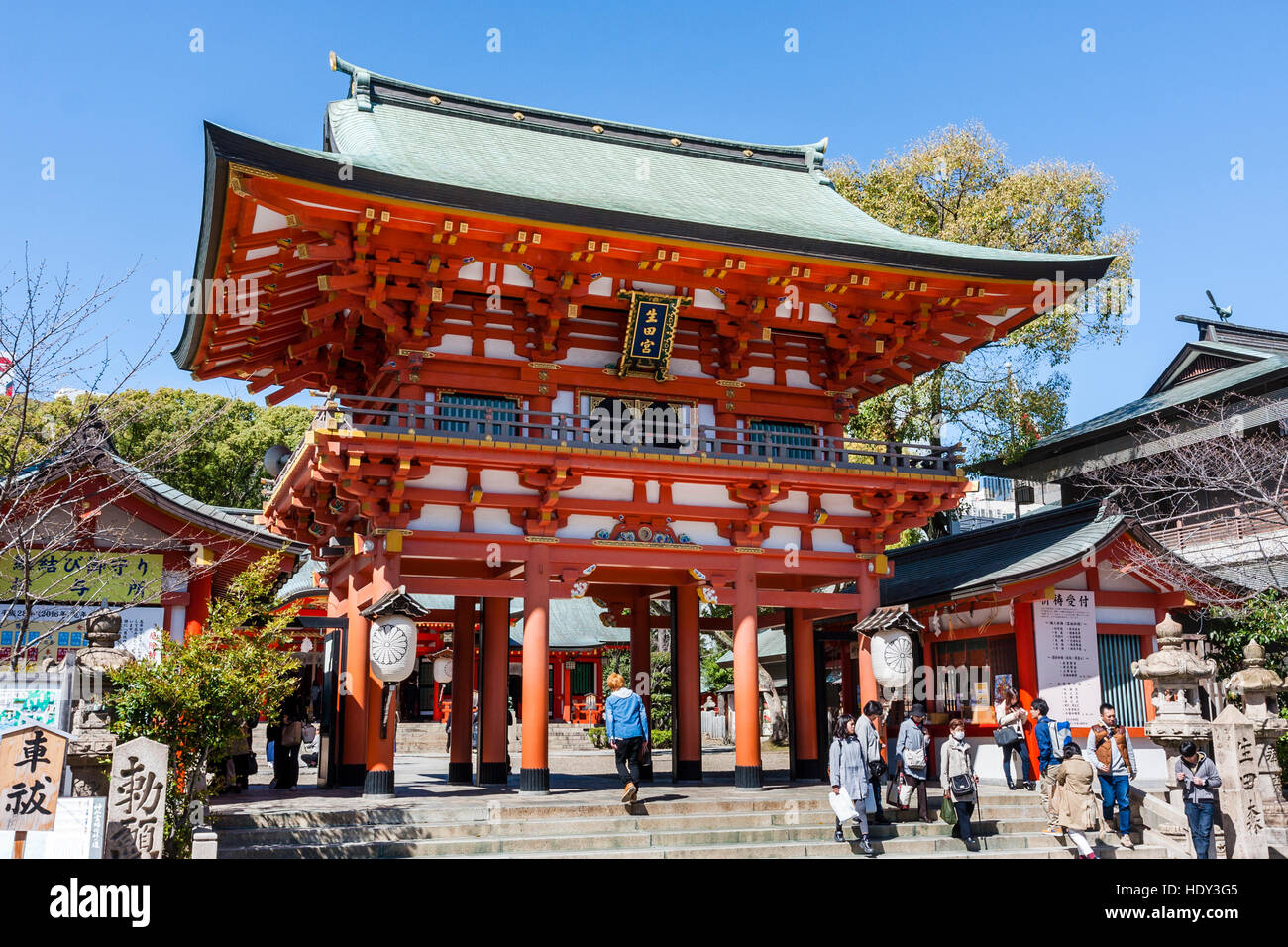 Japan, Kobe, Ikuta shrine. Ikuta-jinja. Two storey vermilion mon, gate, entrance to shrine. People walking through. Bright sunshine, blue sky. Stock Photo