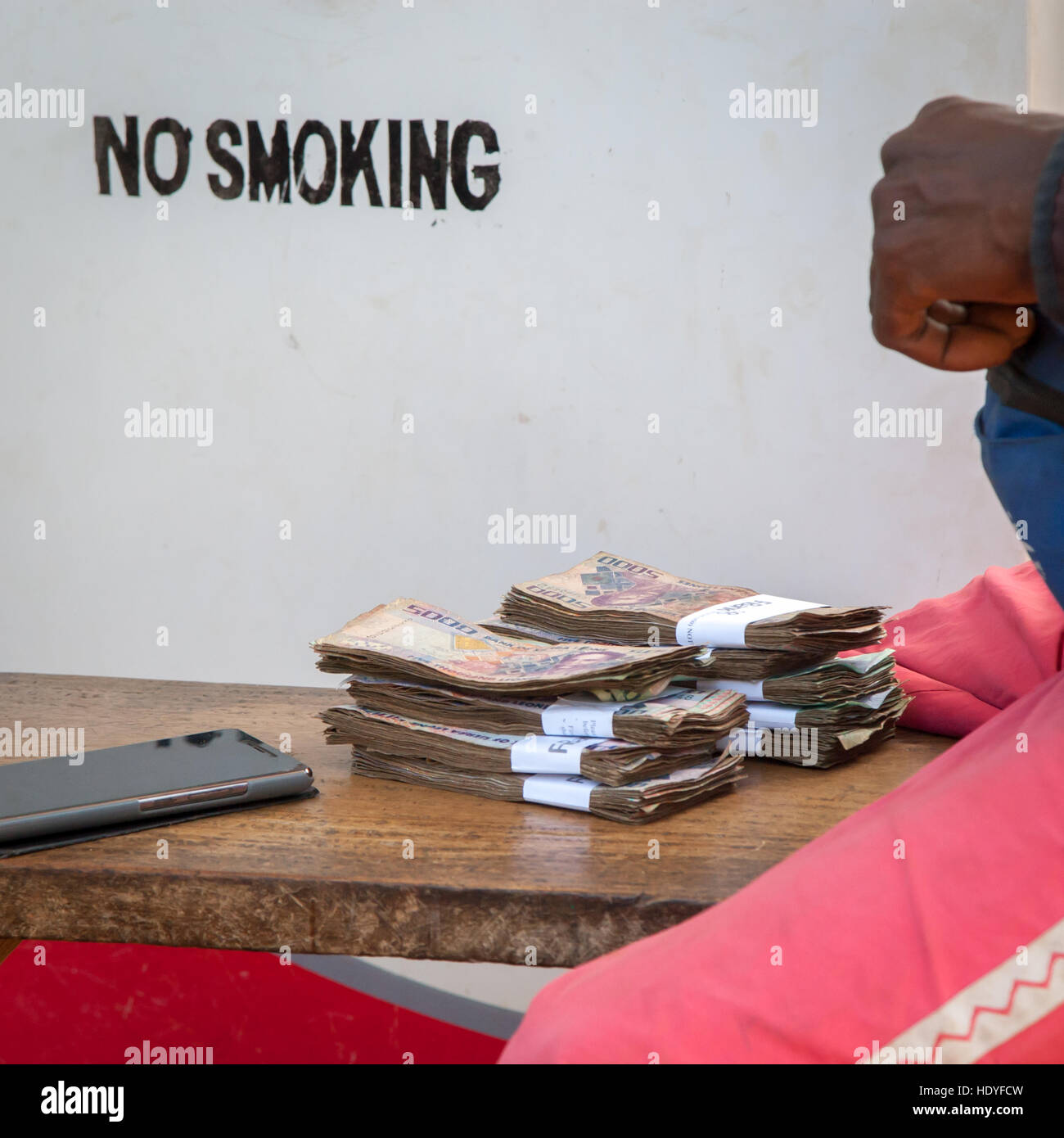 Bundled leone banknotes lie on a wooden table beside a gas station in Tuala, Sierra Leone. A sign behind them prohibits smoking. Stock Photo