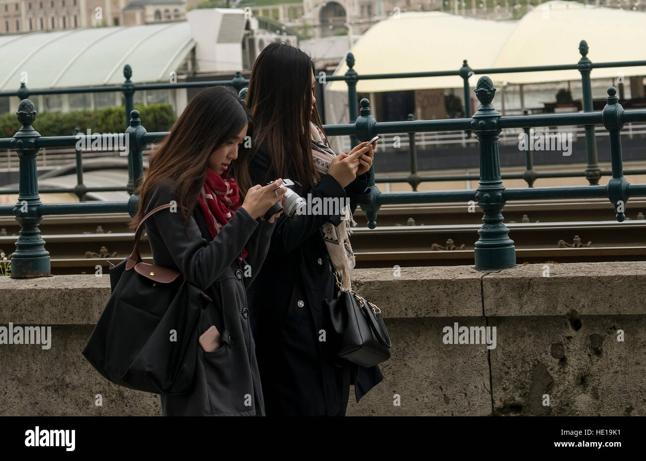 Budapest, Hungary - April 11,2016: Two young Chinese girl walking by the fence and play with mobile phone. Stock Photo