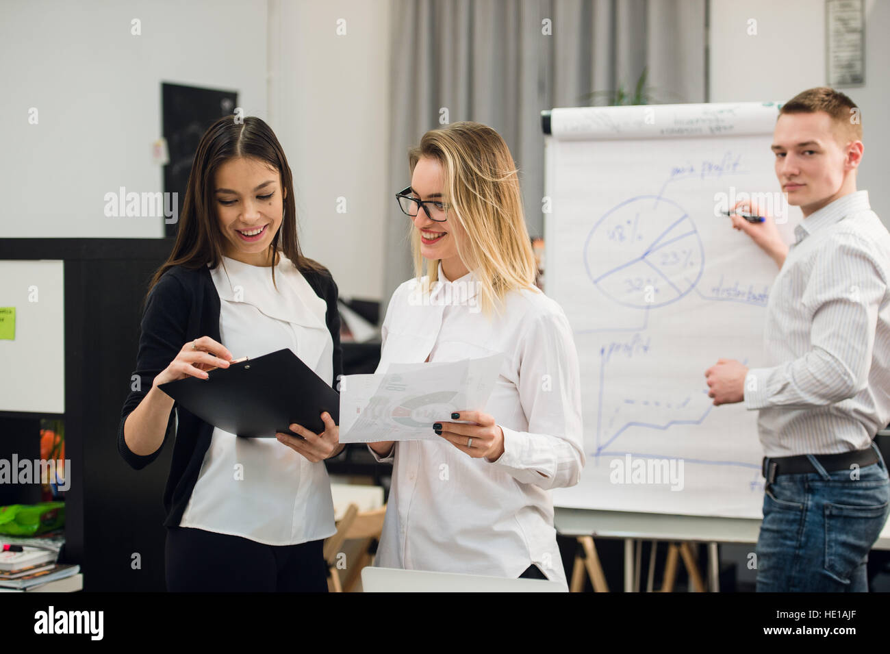 Two beautiful office workers having conversation while man colleague drawing business strategy on flip chart. Stock Photo