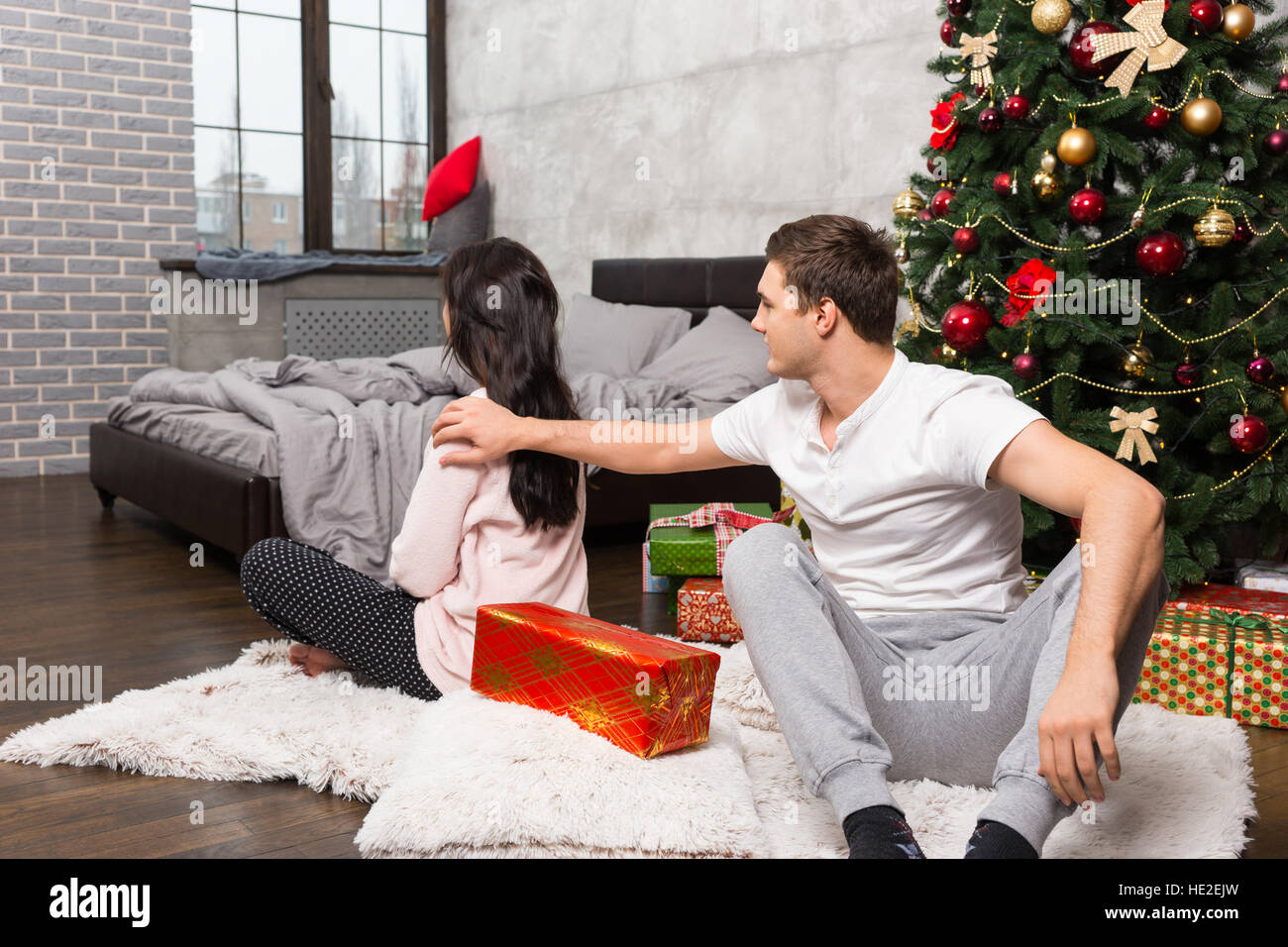 Offended girl upset about the wrong gift of her boyfriend while sitting on the carpet near Christmas tree in the room in loft style Stock Photo