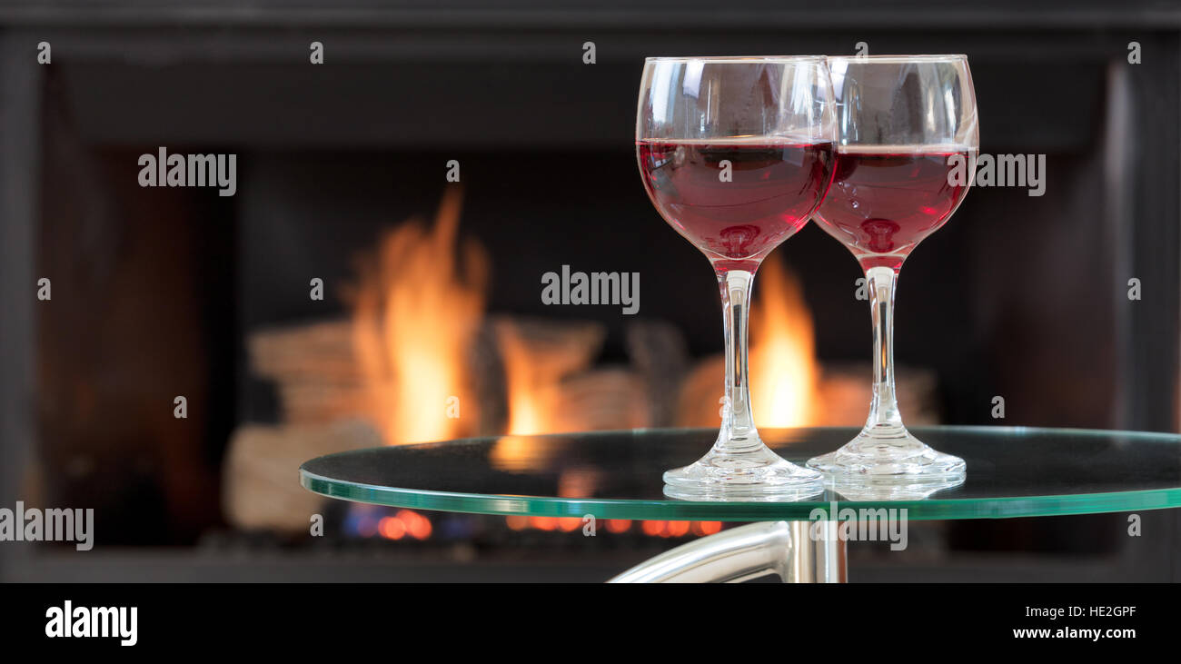 Two glasses of red wine on top of glass table with glowing fireplace in ...