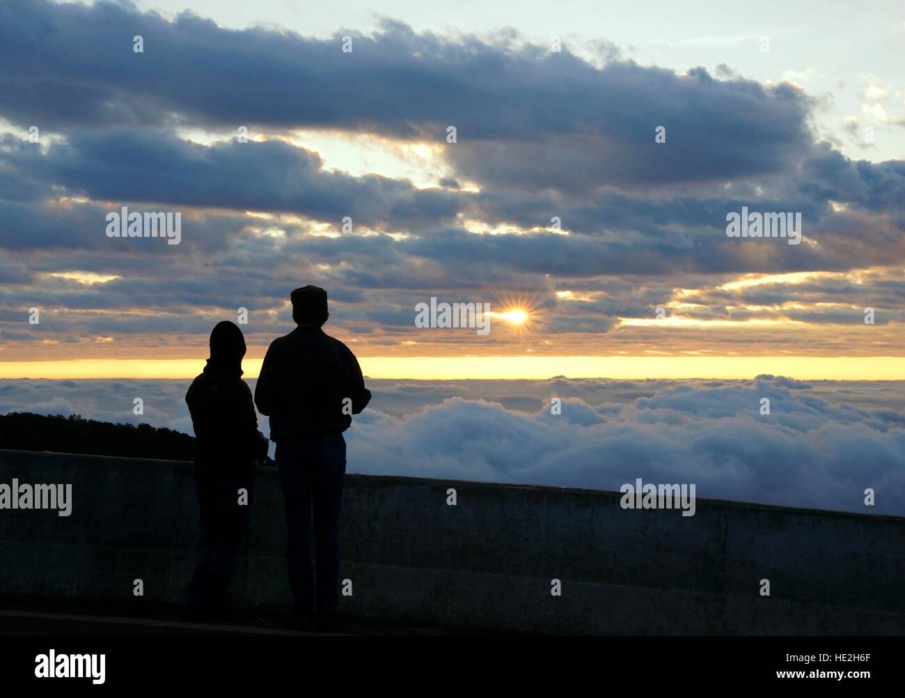 Silhouette couple looking at the beautiful sunrise on the top of mountain with the view into misty valley. Morning mountain landscape with wave of fog Stock Photo