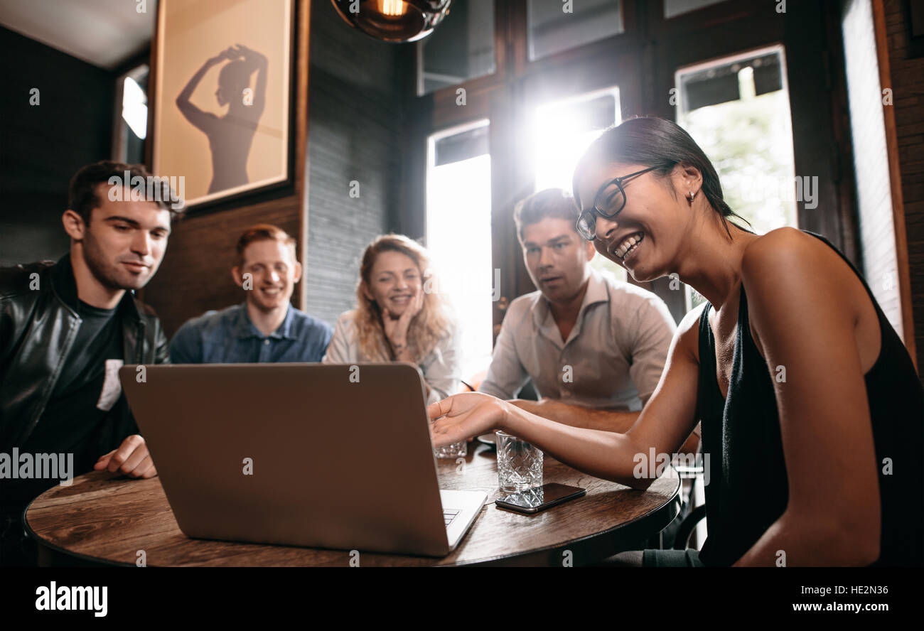 Smiling young woman showing something on laptop to her friends. Group of young men and women at cafe looking at laptop computer and smiling. Stock Photo