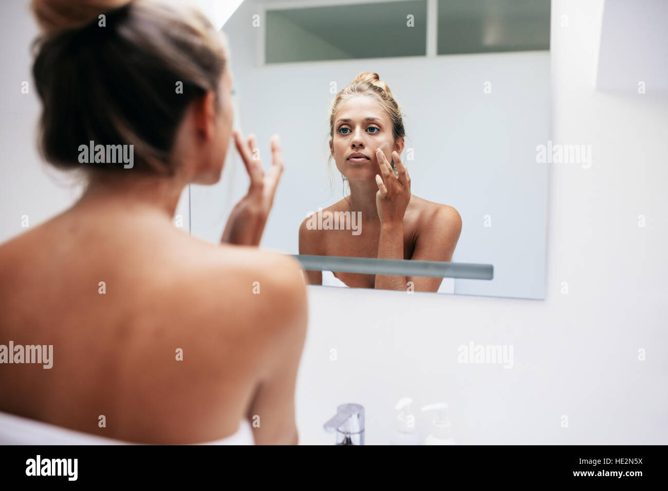 Woman in bathroom looking in to mirror and applying cream on her face . Beautiful female pampering her facial skin. Stock Photo