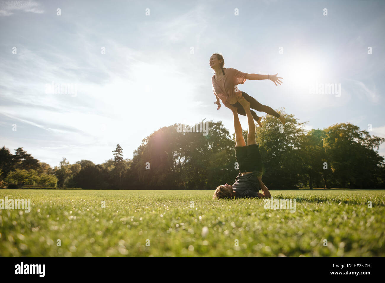 Fit young couple doing acrobatic yoga in park. Man lying on grass and balancing woman in his feet. Stock Photo