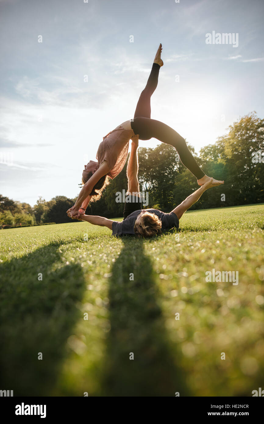Vertical shot of healthy young couple doing acrobatic yoga on grass. Man and woman exercising in pair at the park. Stock Photo