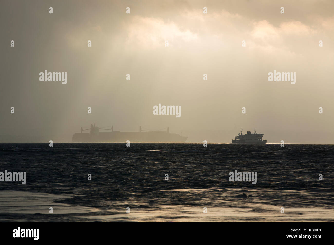 Break in the storm in the Solent. Outlines of a bulk carrier and car ferry on the horizon on this dark day with light breaking. Stock Photo