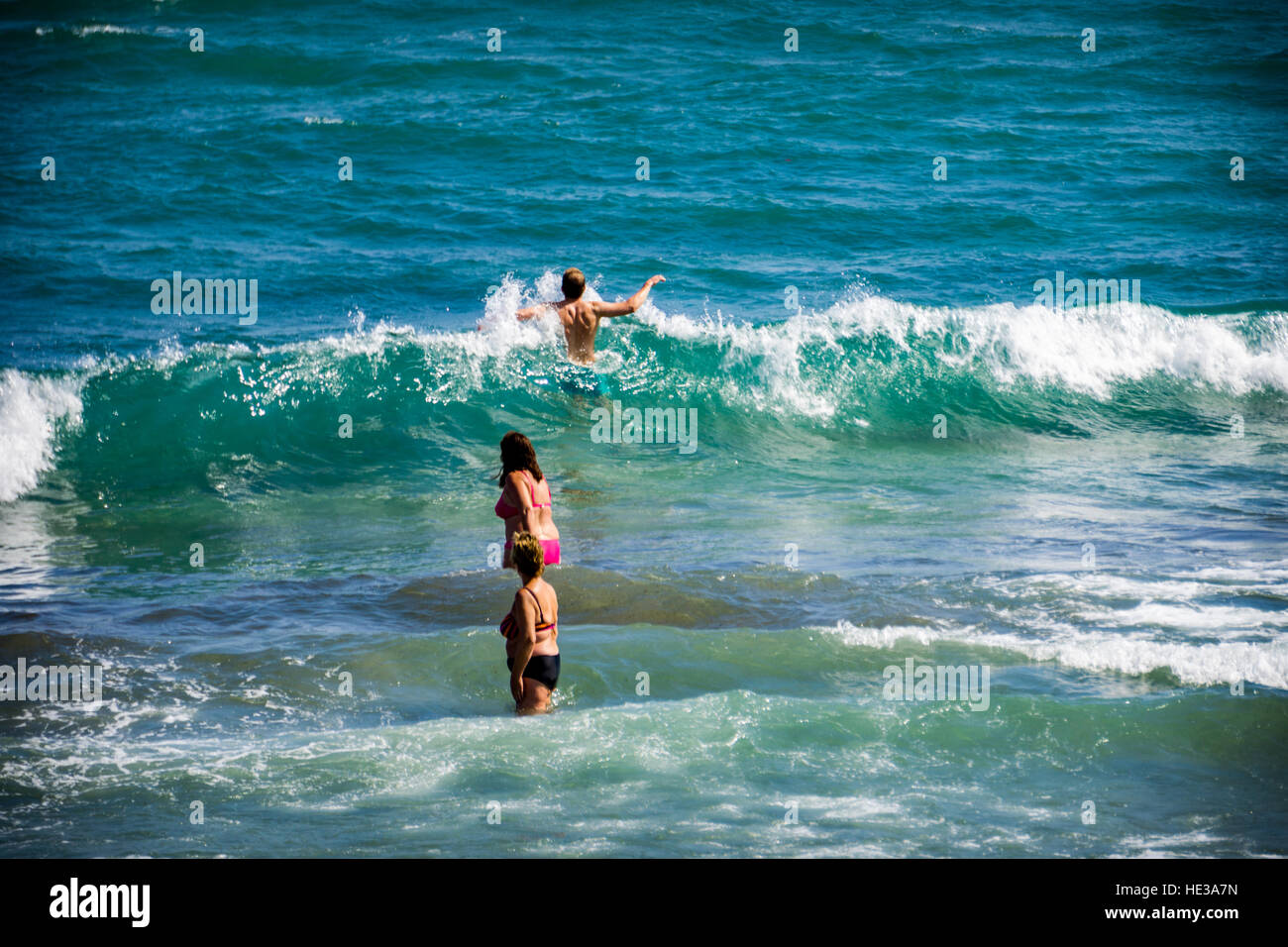 Two middle aged women paddle in the sea while watching a young man jump through the surf Stock Photo