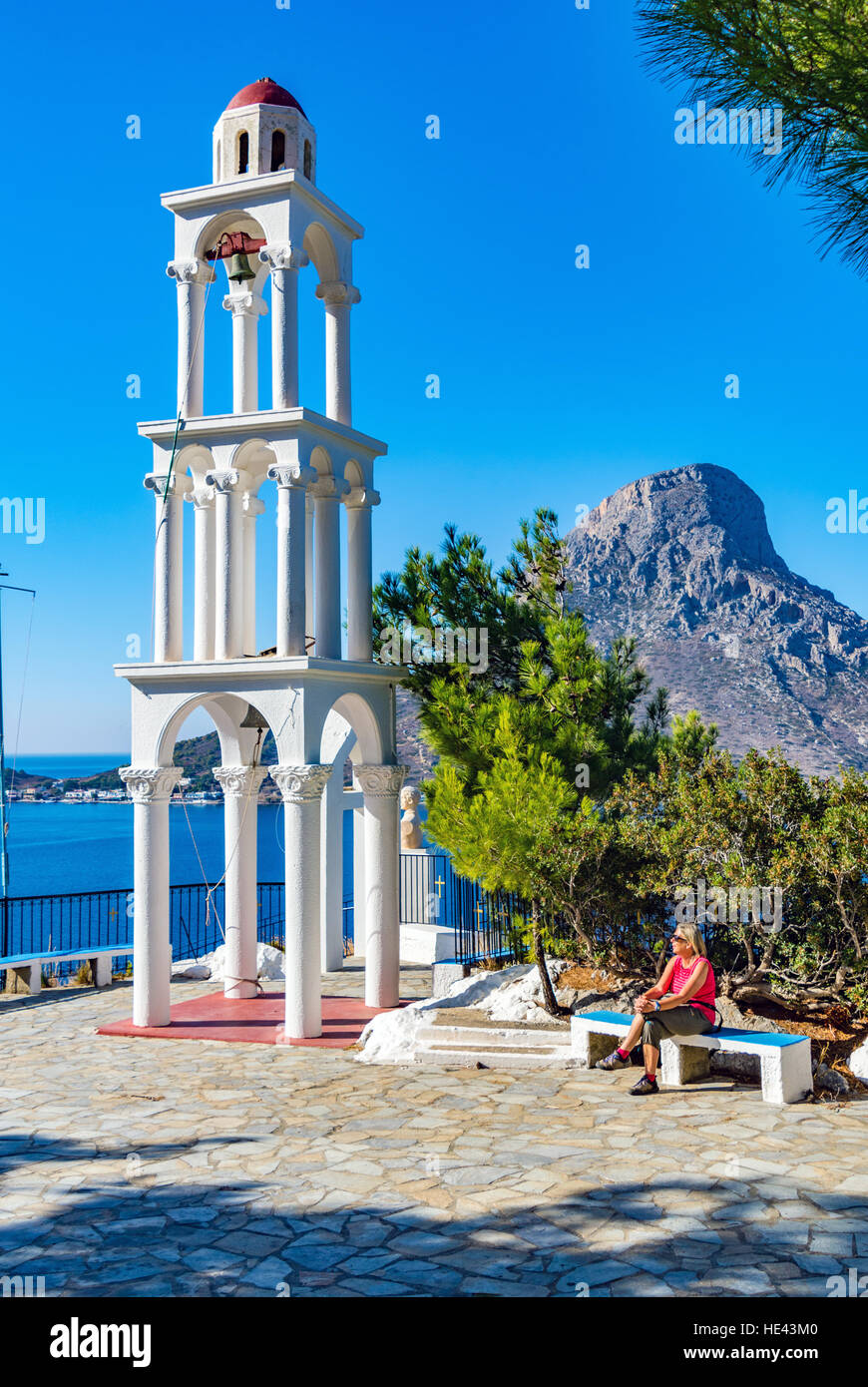 Female figure in red sat by white bell tower, blue sky, blue sea, Kalymnos Stock Photo