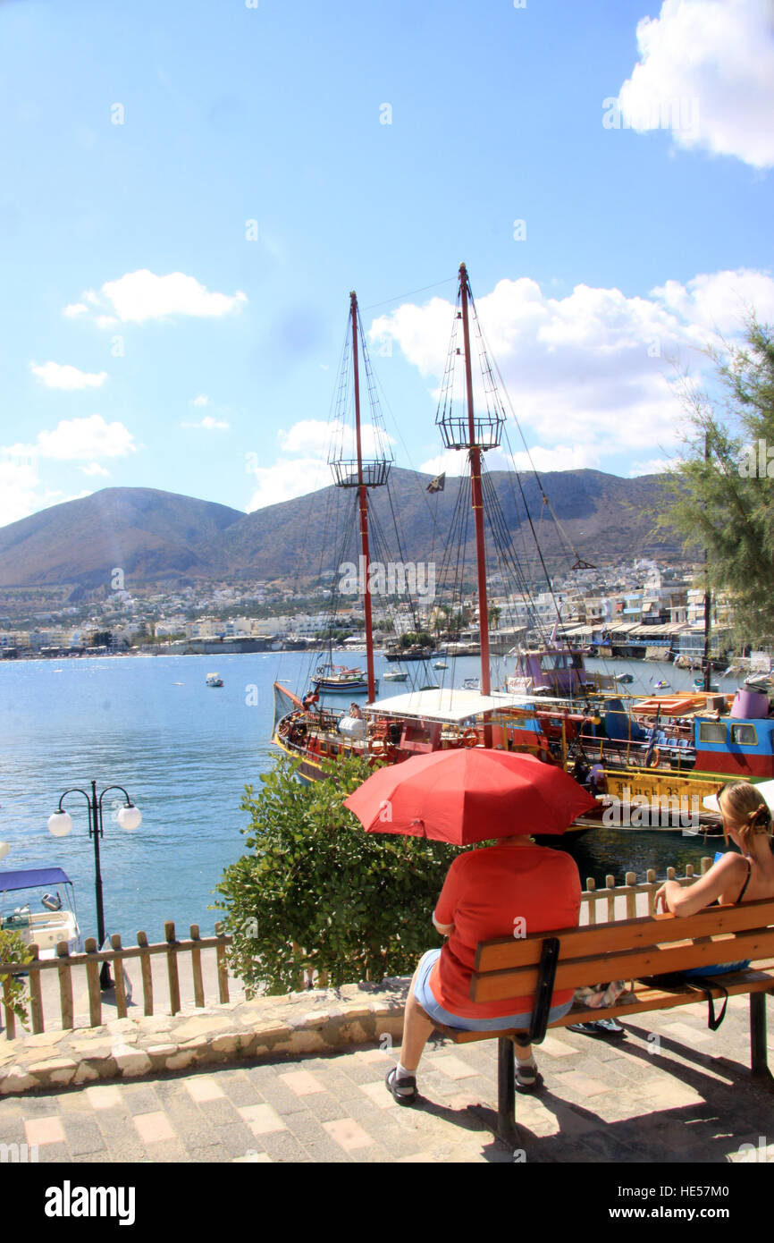 Holiday maker sitting under a red parasol to keep the sun off taking in the view at  Agios Nikolaos on the Greek island of Crete Stock Photo