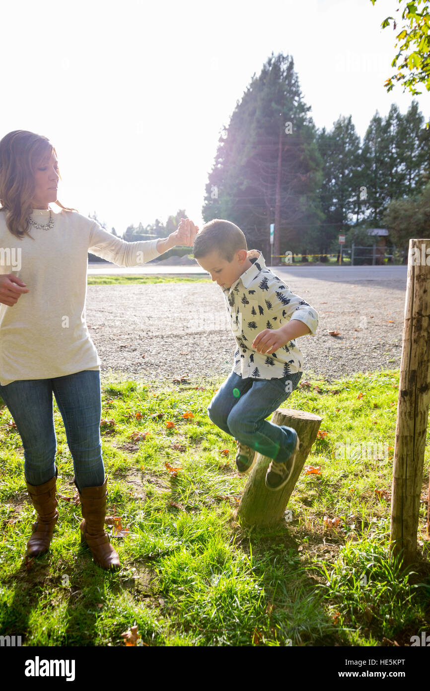 Boy jumping off a wooden post while holding the hand of his mother at a park. Stock Photo