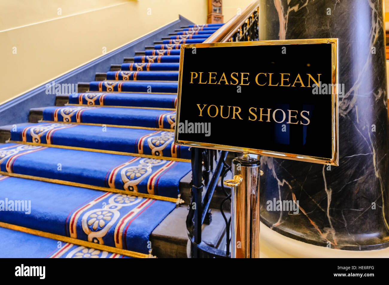Sign saying 'Please clean your shoes' at the bottom of a flight of stairs in a large, ornate building. Stock Photo
