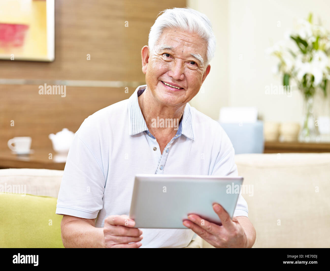 senior asian man sitting on sofa holding a tablet computer looking at camera smiling. Stock Photo