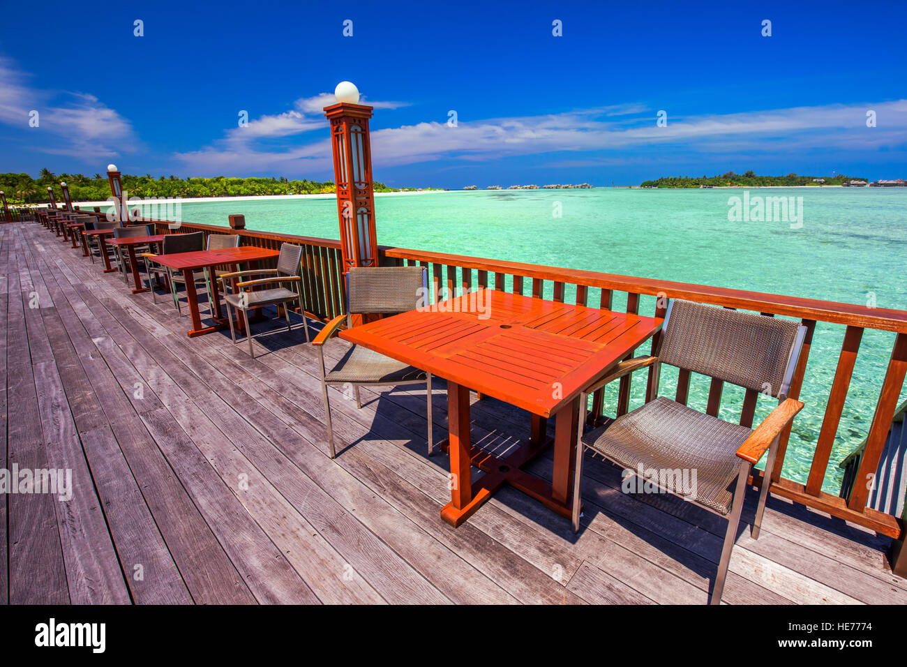 Chairs and tables with palm trees, water bungalows and clear water on luxurious tropical beach resort. Stock Photo