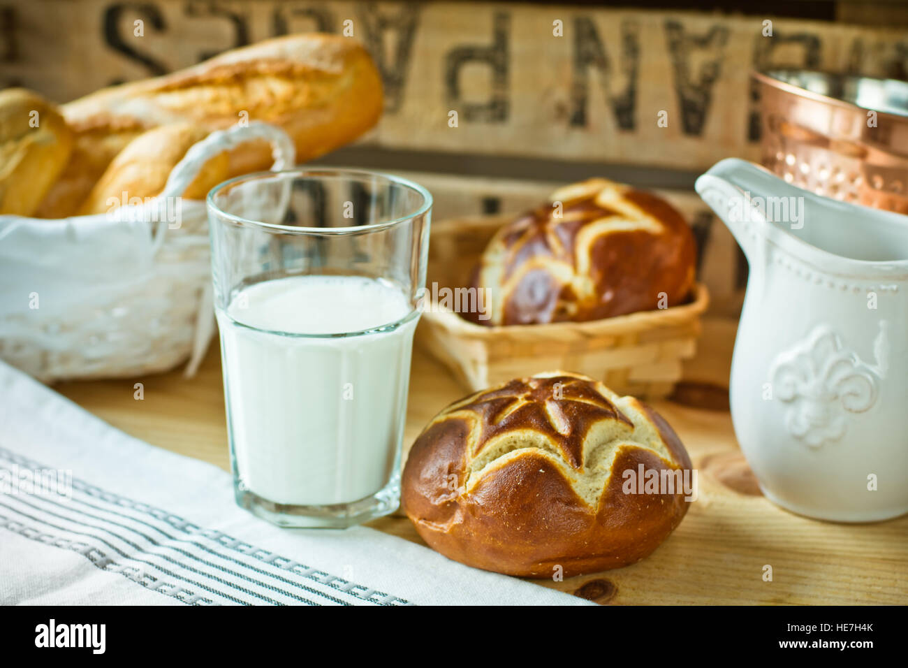 Fresh lye rolls in a wicker basket, a glass of milk, white pitcher on a wood table in a rustic style kitchen interior Stock Photo
