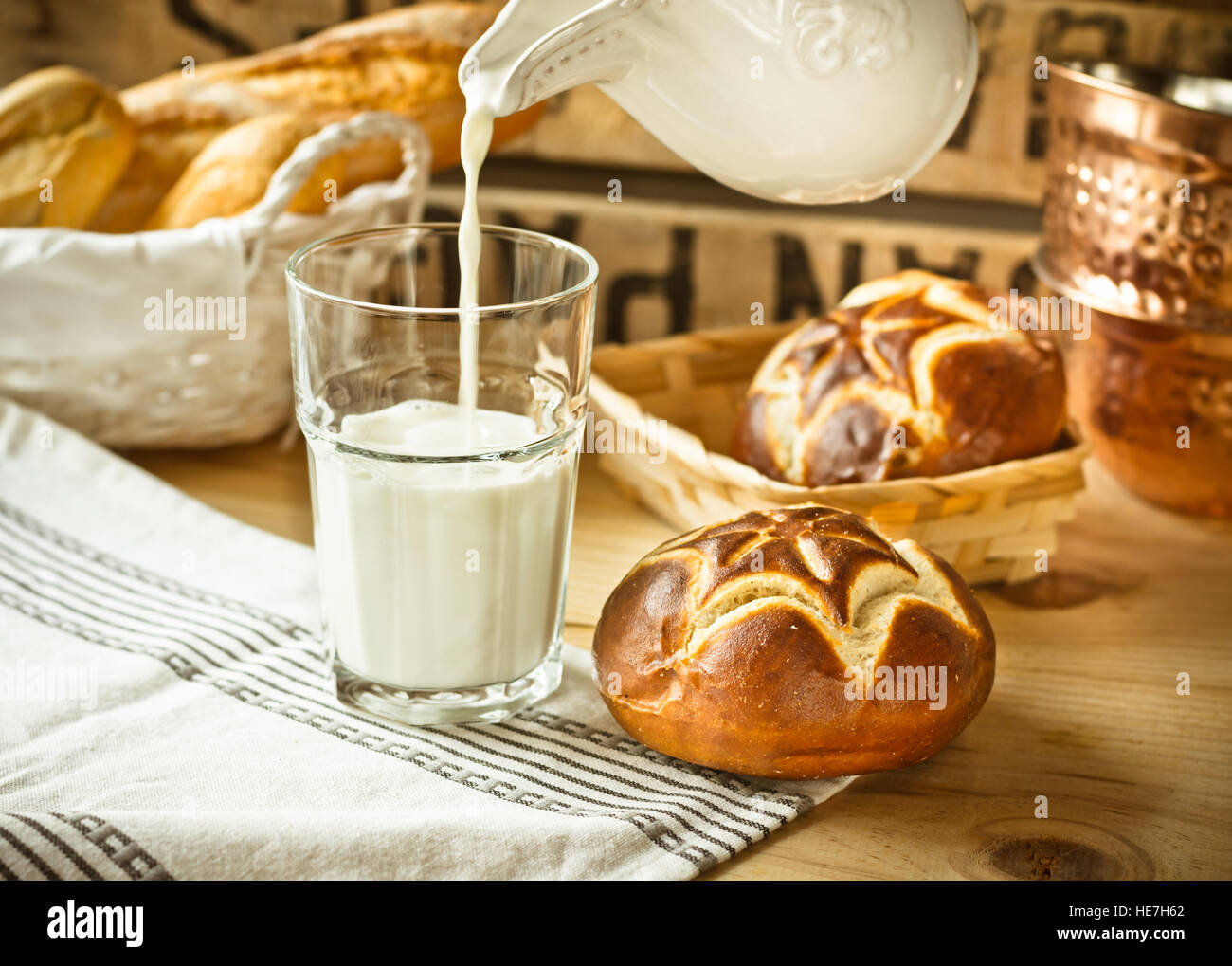 Fresh lye rolls in a wicker basket, process of pouring milk into a glass from pitcher, wood table, rustic style kitchen interior Stock Photo