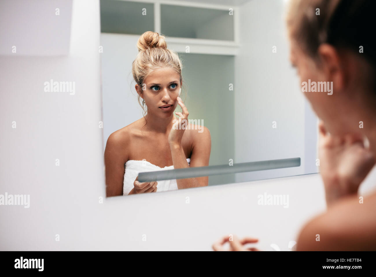 Beautiful young woman applying cosmetic cream on face standing in the bathroom. Reflection in female doing skin treatment. Stock Photo