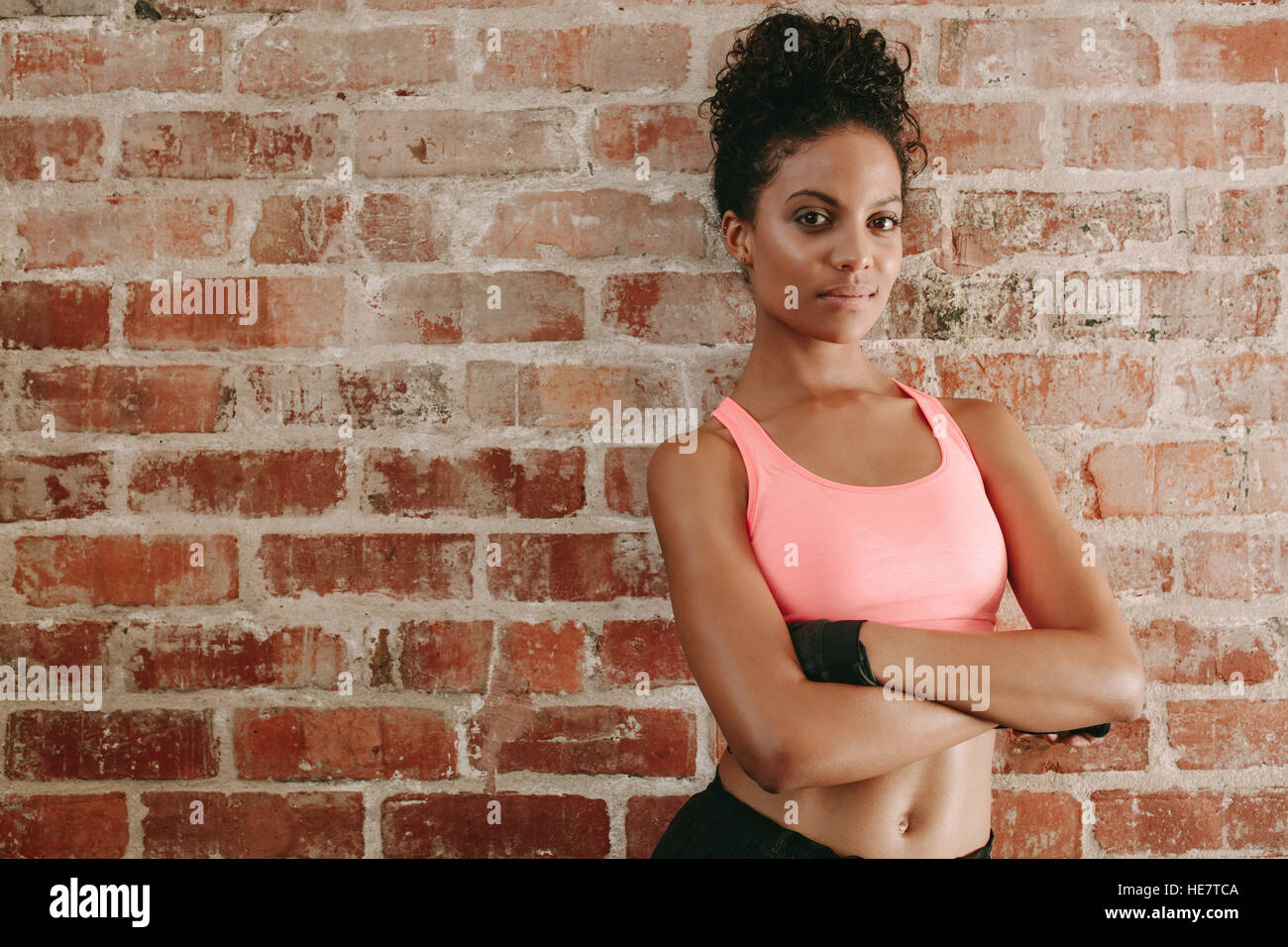 Portrait of young african woman in sports bra standing against brick wall with her arms crossed. Female fitness model in gym. Stock Photo